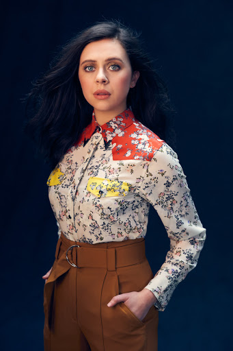 Portrait of a woman with dark wavy hair wearing a floral print shirt and brown high waisted trousers against a dark textured background