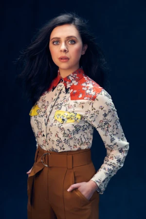 Portrait of a woman with dark wavy hair wearing a floral print shirt and brown high waisted trousers against a dark textured background