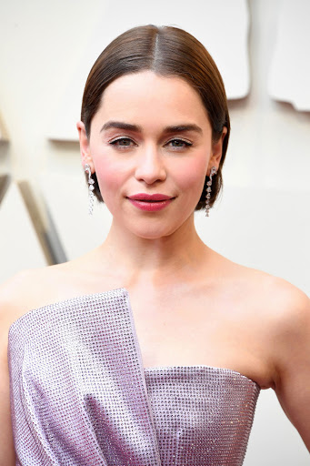 Close-up portrait of Emilia Clarke on red carpet with soft lighting, wearing shiny lilac dress and long dangling diamond earrings.
