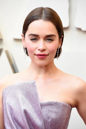 Close-up portrait of Emilia Clarke on red carpet with soft lighting, wearing shiny lilac dress and long dangling diamond earrings.
