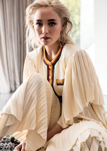 Blonde woman in cream-colored pleated jumpsuit with buttoned jacket poses indoors with soft lighting and a neutral background.