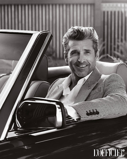 Black and white portrait of a smiling mature man in a houndstooth blazer in a vintage convertible, outdoors, with L'Officiel logo.