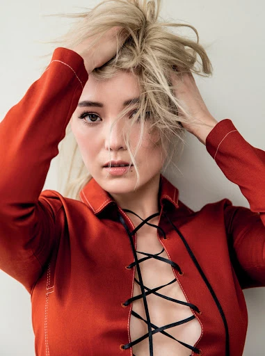 Close-up portrait of a blonde woman in a rust-colored shirt with lace-up detail, posing against a neutral background, hands in her hair.
