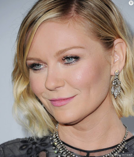 Close-up portrait of Kirsten Dunst wearing ornate dangling earrings and a beaded necklace against a plain backdrop.