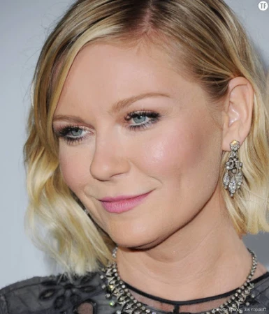 Close-up portrait of Kirsten Dunst wearing ornate dangling earrings and a beaded necklace against a plain backdrop.