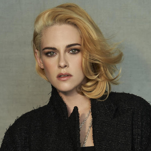 Close-up studio portrait of a blonde woman in a dark coat, with short wavy hair, looking at the camera against a grey background.