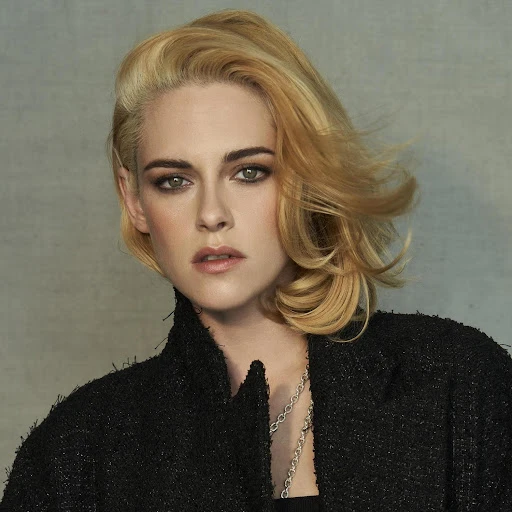 Close-up studio portrait of a blonde woman in a dark coat, with short wavy hair, looking at the camera against a grey background.