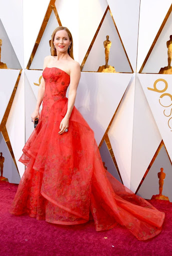 Woman in coral strapless gown on red carpet at awards show with white triangular patterned background, portrait orientation.