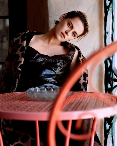Indoor portrait of a woman with short blonde hair, wearing a black dress and leopard print cardigan, sitting at a pink metal table.