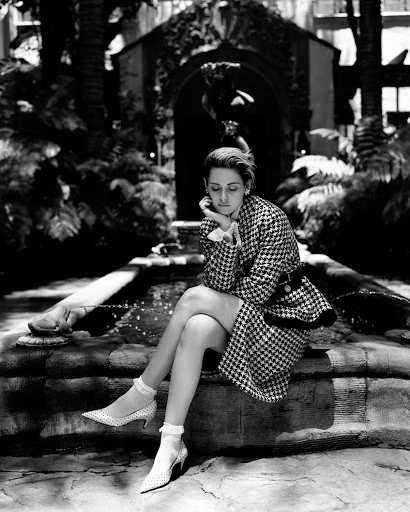 Black and white outdoor shot of a woman in a houndstooth suit sitting on stone steps near a fountain, exuding elegance and classic style.