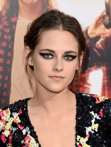 Close-up of Kristen Stewart with heavy black eye makeup and a shimmering black V-neck dress posing on a red carpet at a premiere event.