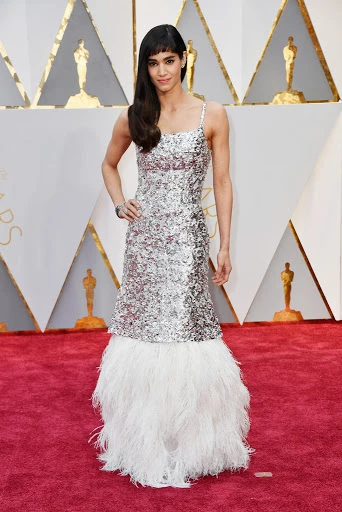 Elegant woman in sequin gown with feathered hem stands on a red carpet with golden statues in the backdrop, at a formal event.