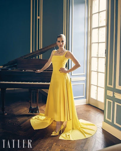 Emma Roberts poses in a yellow dress with a long train, standing next to a piano, in a high-fashion editorial photography shoot.