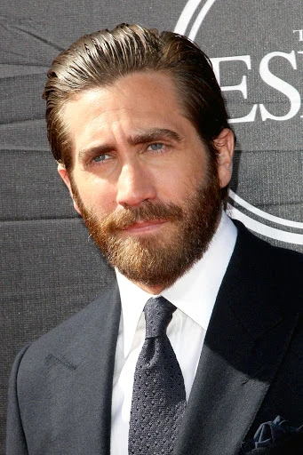 Close-up portrait of Jake Gyllenhaal at an event. He wears a dark suit, white shirt, and patterned tie, standing against a gray backdrop.
