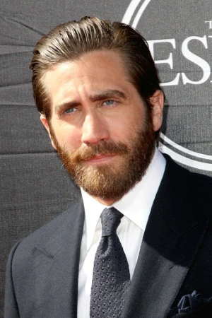 Close-up portrait of Jake Gyllenhaal at an event. He wears a dark suit, white shirt, and patterned tie, standing against a gray backdrop.