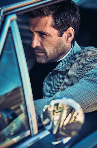 Adult male in a plaid blazer sits inside of a blue car looking out of the open window with the side mirror in view.