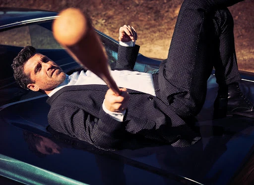 Adult male in a suit and boots leans on a car, holding a baseball bat in an outdoor shot with blurred earthy-toned background.