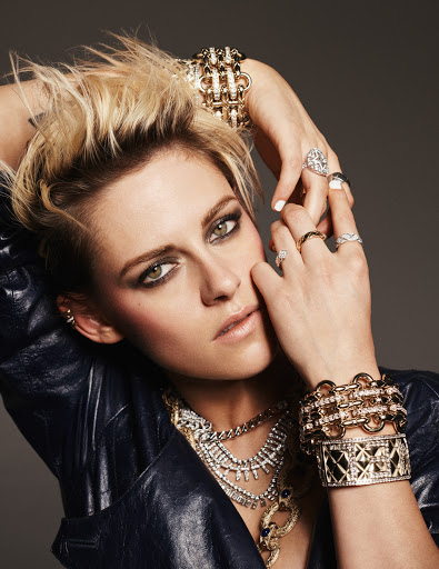 Close-up portrait of a fair-skinned woman with short blonde hair wearing a black jacket and gold and silver jewelry against a grey backdrop.