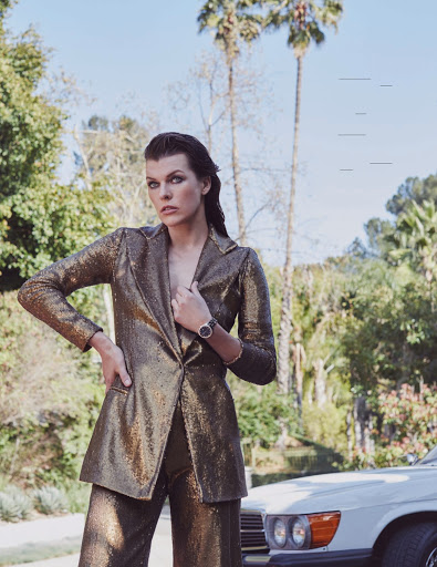 An adult female model poses outdoors in a sequined pantsuit in front of a white car and palm trees against a bright sky.