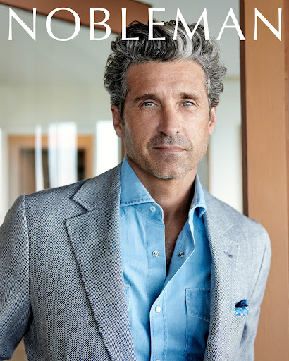 Mature man with grey hair in a blue shirt and grey blazer poses indoors in a portrait shot. Man gazes at camera for magazine cover.