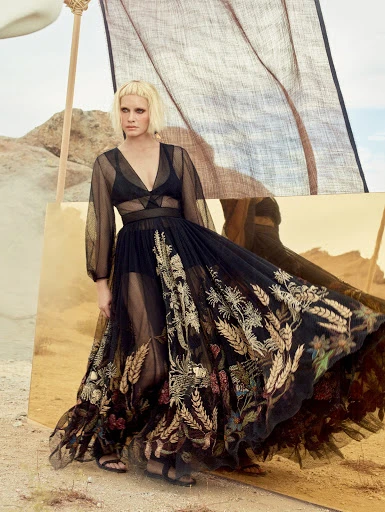 Woman in a sheer black embroidered dress poses in a desert landscape with golden mirrored panels, creating a surreal, fashionable scene.
