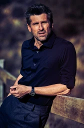 Portrait of actor Patrick Dempsey leaning against a wooden fence wearing a dark button-down shirt and dark trousers with a wristwatch.
