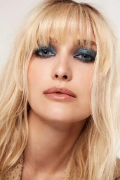 Close-up portrait of a blonde woman with heavy blue smokey eye makeup and textured fringe bangs against a neutral background.