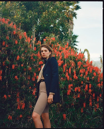 Woman poses in front of red flowers wearing a blazer jacket, tan top, and cycling shorts in a natural outdoor environment with a palm tree.