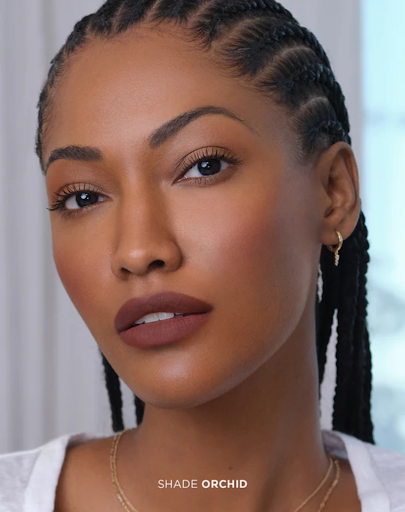 Close-up of an adult woman with braids, wearing matte lipstick, jewelry, and a neutral top. The background is out of focus.