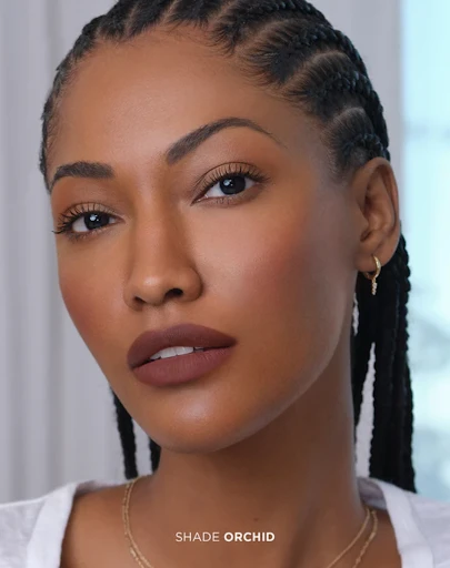 Close-up of an adult woman with braids, wearing matte lipstick, jewelry, and a neutral top. The background is out of focus.