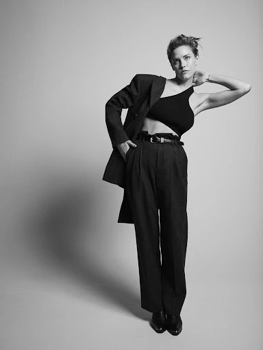 Woman in a black one-shoulder crop top, dark oversized suit jacket, high-waisted trousers, and black shoes poses in a studio setting.