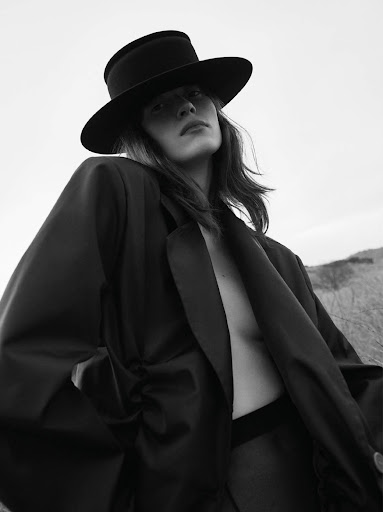 Low-angle black and white portrait of a woman wearing a wide-brimmed hat and an oversized dark jacket outdoors.