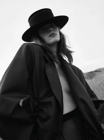Low-angle black and white portrait of a woman wearing a wide-brimmed hat and an oversized dark jacket outdoors.