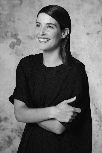 Portrait of an adult woman smiling in a black dress against a mottled grey background, shot in black and white with natural lighting.