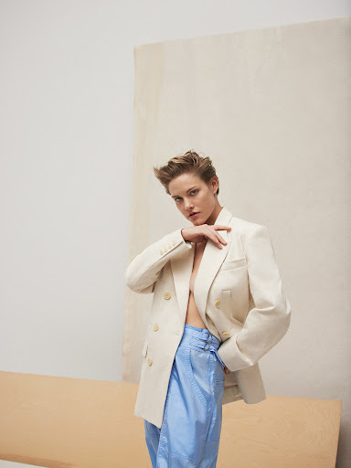 Woman in light-colored blazer and iridescent blue pants strikes a confident pose against a neutral backdrop in a fashion editorial shot.