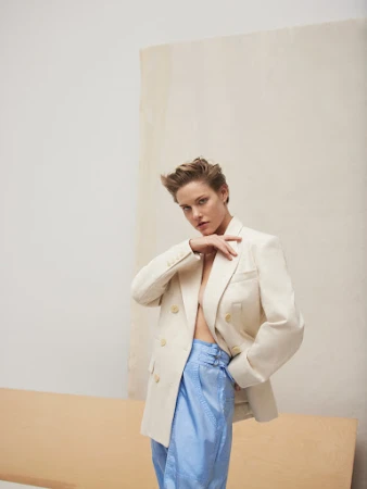 Woman in light-colored blazer and iridescent blue pants strikes a confident pose against a neutral backdrop in a fashion editorial shot.