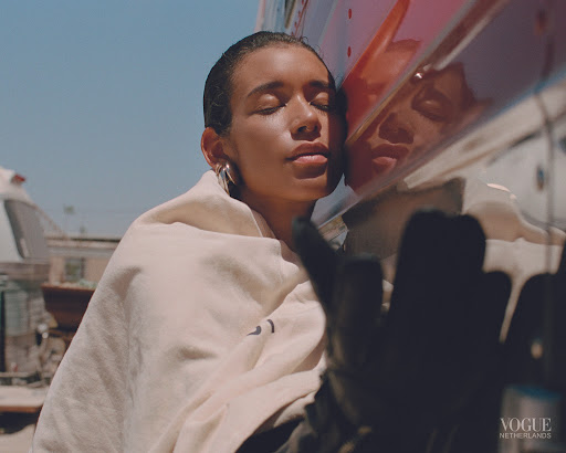 Young woman with eyes closed leans against an orange bus wearing a cream-colored hooded jacket in outdoor daytime fashion shot.