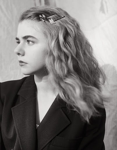 Monochrome portrait of a young woman with wavy hair, wearing a black blazer and a hair clip, against a softly textured background.