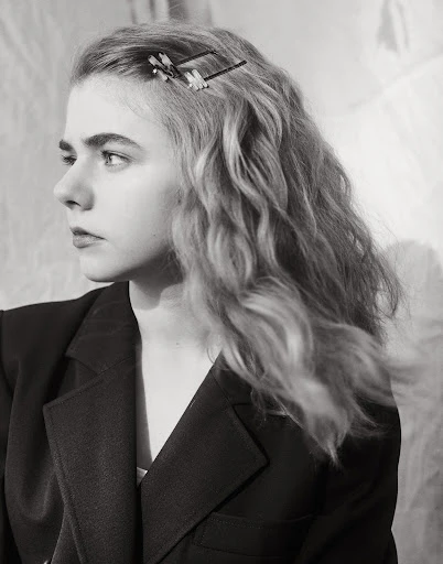Monochrome portrait of a young woman with wavy hair, wearing a black blazer and a hair clip, against a softly textured background.