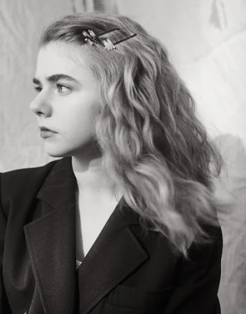 Monochrome portrait of a young woman with wavy hair, wearing a black blazer and a hair clip, against a softly textured background.