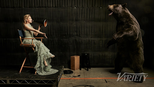 An adult woman in a dress sits in a director's chair with a bullhorn facing a standing bear in a photo studio with dark backdrop.