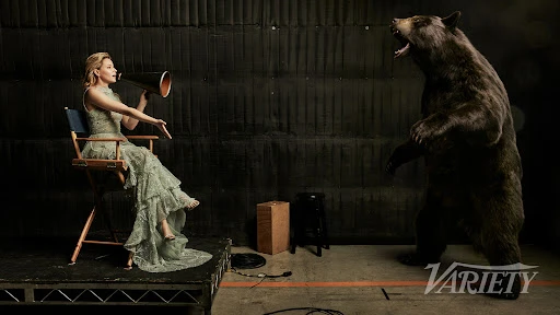 An adult woman in a dress sits in a director's chair with a bullhorn facing a standing bear in a photo studio with dark backdrop.