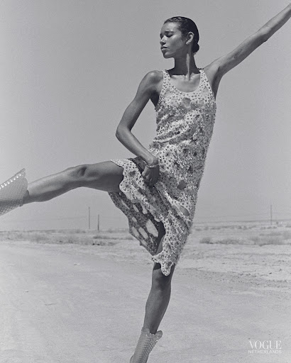 Woman in patterned dress and studded boots strikes a ballet pose in black and white on an outdoor barren landscape with clear sky.