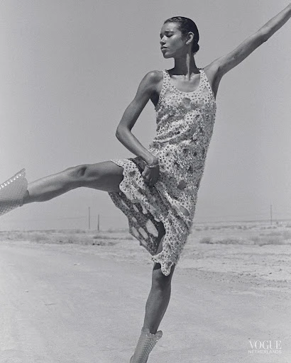 Woman in patterned dress and studded boots strikes a ballet pose in black and white on an outdoor barren landscape with clear sky.