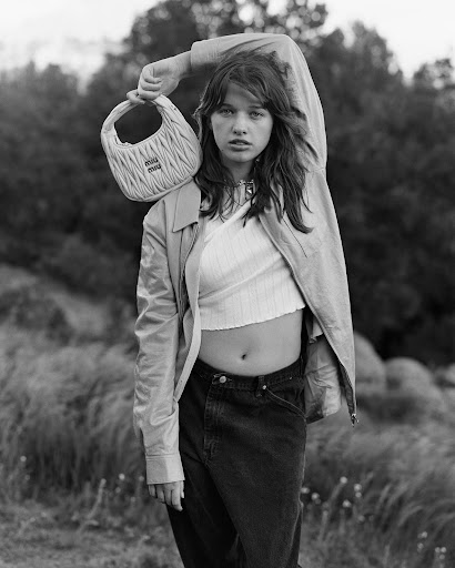 Monochrome fashion shot of a young woman in a crop top and jeans, holding a Miu Miu handbag with soft focus foliage background.