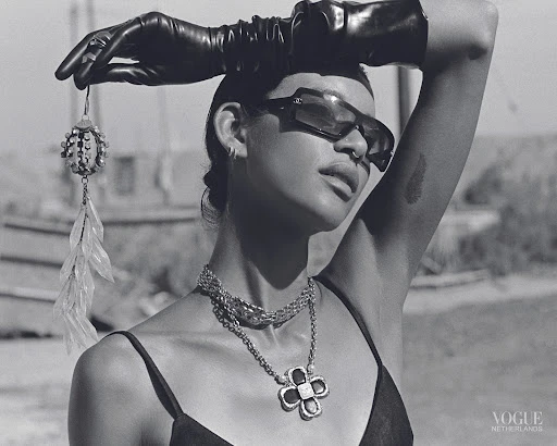 Fashion shot of a woman in sunglasses and gloves holding an earring, with a layered necklace, against an outdoor landscape in black and white.