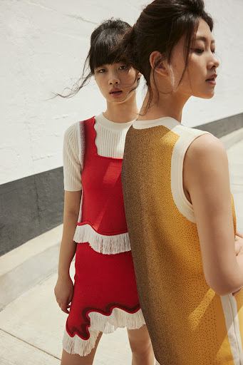 Two Asian women in stylish knitwear stand outdoors, one in a red dress with fringe, the other in a patterned yellow top.