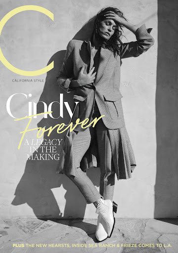 Monochrome magazine cover portrait of a woman leaning against a stone wall in a patterned pantsuit and white ankle boots.