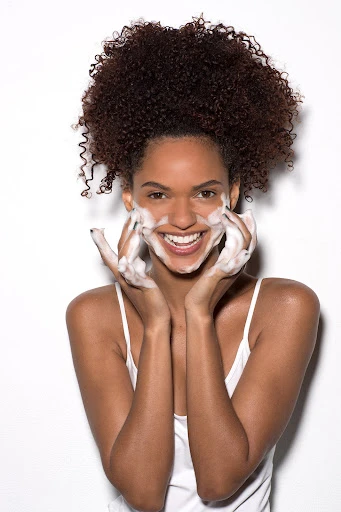 Smiling woman with dark curly hair washing her face with cleanser in a close-up studio shot with white background, beauty concept.