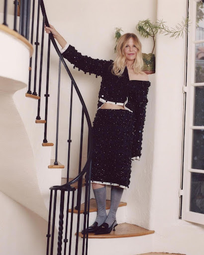 Woman in a black sequined skirt and jacket poses on a white spiral staircase indoors, near a window and potted plant, for a fashion shot.